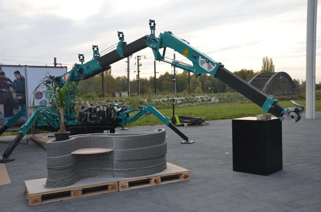 Robotic arm 3D concrete printer creating a curved bench-shaped wall outdoors